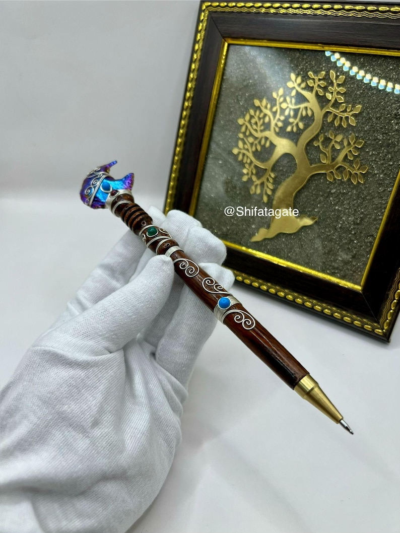 May include: A brown wooden pen featuring silver wire filigree, small green and blue inlaid stones, and a metallic gold tip. The top of the pen is adorned with a faceted, iridescent blue and purple crystal. The text @Shifatagate is visible.
