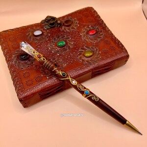 May include: A dark brown pen with gold accents and colorful gemstones. The pen is lying on a brown leather journal with embossed floral designs and colorful gemstones. The pen has a crystal at the top and the text @SHIFATAGATE.