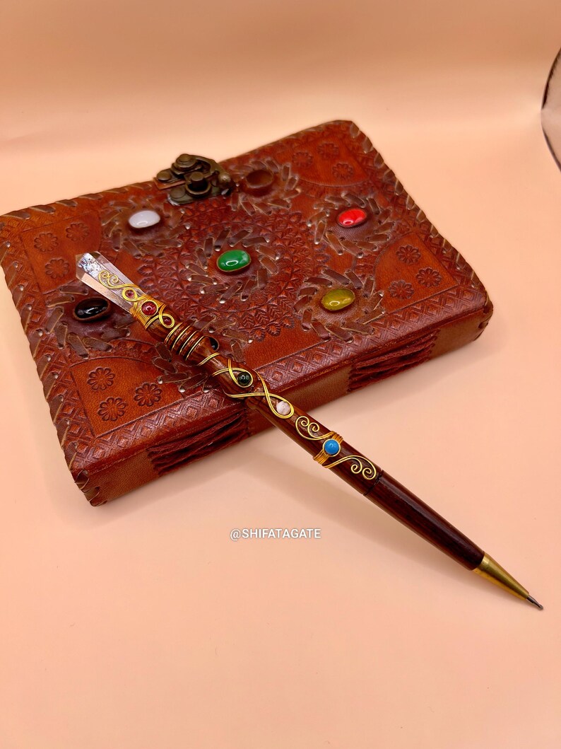 May include: A dark brown pen with gold accents and colorful gemstones. The pen is lying on a brown leather journal with embossed floral designs and colorful gemstones. The pen has a crystal at the top and the text @SHIFATAGATE.