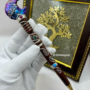 May include: A handcrafted pen with a dark wood barrel and a decorative top. The top features a crescent moon shape with iridescent blue and purple accents, silver wire detailing, and gemstone embellishments. The pen is held in a gloved hand.