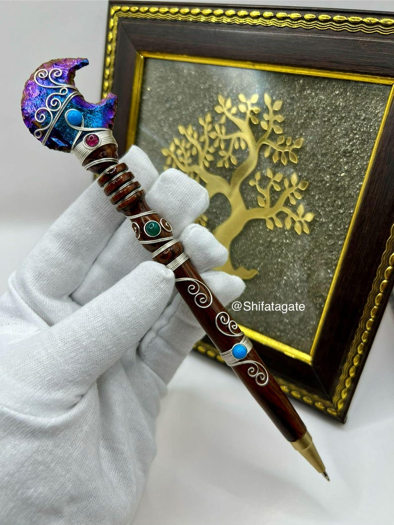 May include: A handcrafted pen with a dark wood barrel and a decorative top. The top features a crescent moon shape with iridescent blue and purple accents, silver wire detailing, and gemstone embellishments. The pen is held in a gloved hand.