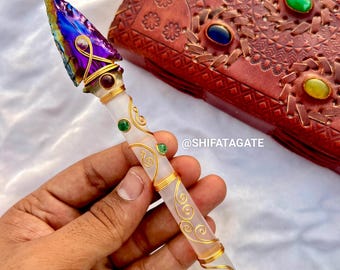 May include: A handcrafted crystal wand with a multi-colored, iridescent arrowhead tip. The wand features gold wire accents, a clear handle, and small gemstones. The text "@SHIFATAGATE" is visible.