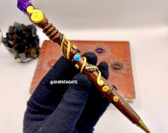 May include: A handcrafted wooden pen with a purple crystal top and gold accents. The pen features a dark brown finish, a turquoise stone, and a brass tip. The pen is held by a person wearing a black glove. The text "@SHIFATAGATE" is visible.