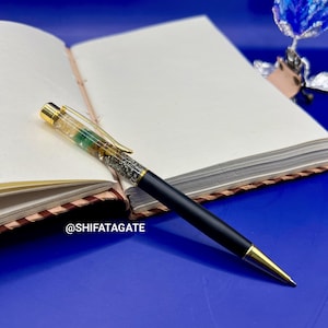 K&ouml;nnte beinhalten: Ein schwarzer Stift mit goldenen Akzenten und einem dekorativen Aufsatz, der auf einem offenen Notizbuch liegt. Der Stift hat klare und gr&uuml;ne Elemente. Der Text "@SHIFATAGATE" ist sichtbar.