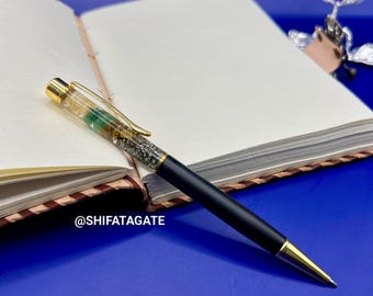 May include: A black pen with gold accents and a decorative top, resting on an open notebook. The pen's top features a mix of clear and green elements. The text "@SHIFATAGATE" is visible.