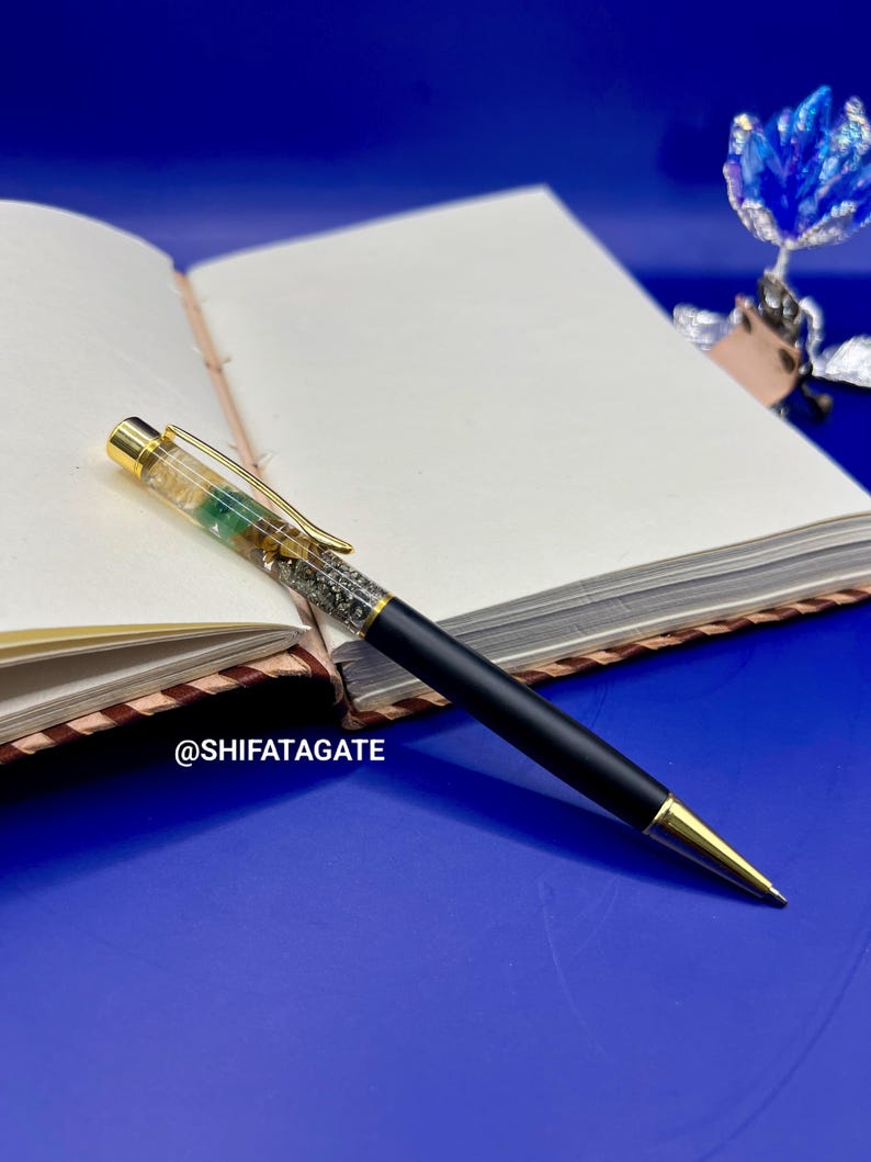 K&ouml;nnte beinhalten: Ein schwarzer Stift mit goldenen Akzenten und einem dekorativen Aufsatz, der auf einem offenen Notizbuch liegt. Der Stift hat klare und gr&uuml;ne Elemente. Der Text "@SHIFATAGATE" ist sichtbar.