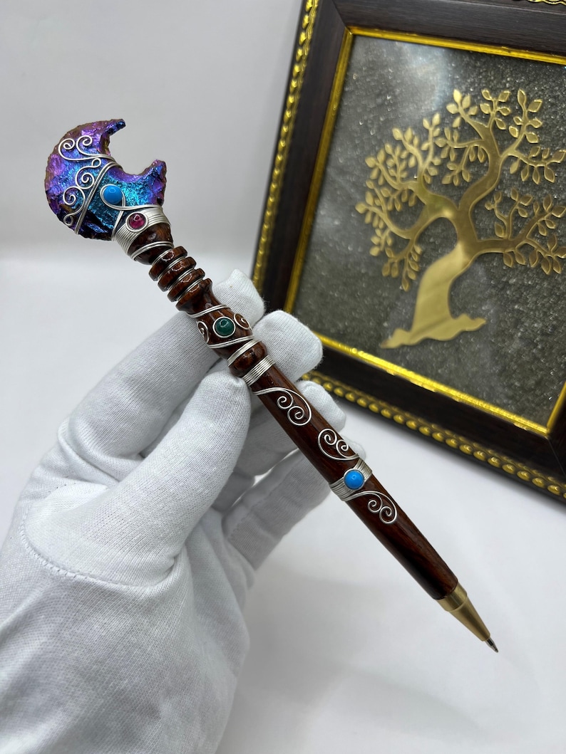 May include: A handcrafted pen with a dark wood barrel and a decorative top. The top features a crescent moon design with iridescent purple and blue hues, accented with silver wire and gemstones. The pen is held by a gloved hand.