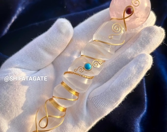 May include: A crystal wand with a pink rose quartz sphere at the top, wrapped in gold wire with turquoise and red gemstone accents. The wand is held in a white gloved hand.