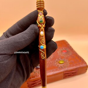 May include: A handcrafted wooden pen with gold accents and colorful gemstones. The pen features a dark brown finish, intricate gold wire detailing, and is adorned with red, green, blue, and clear stones. The pen's tip is gold-colored.