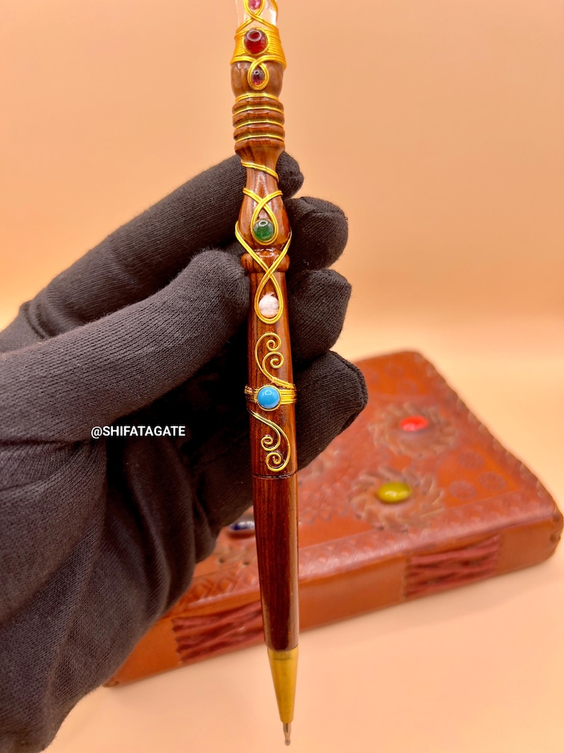 May include: A handcrafted wooden pen with gold accents and colorful gemstones. The pen features a dark brown finish, intricate gold wire detailing, and is adorned with red, green, blue, and clear stones. The pen's tip is gold-colored.