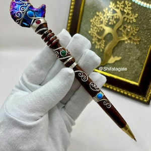 May include: A handcrafted pen with a dark wooden barrel and ornate silver accents. The pen features a decorative top with a crescent moon design, adorned with iridescent colors and gemstones. The pen's design includes turquoise and red gemstones.