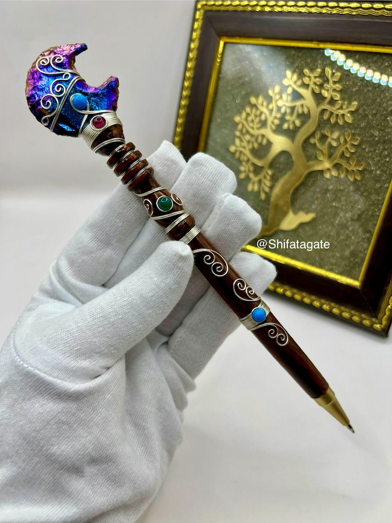 May include: A handcrafted pen with a dark wooden barrel and ornate silver accents. The pen features a decorative top with a crescent moon design, adorned with iridescent colors and gemstones. The pen's design includes turquoise and red gemstones.