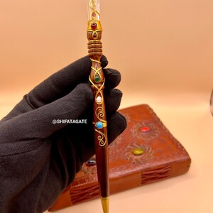 May include: A decorative pen with a clear crystal top and gold accents. The pen features a dark brown wooden body adorned with red, green, and turquoise gemstones. The pen's design includes gold wire wrapping and a gold-colored tip.