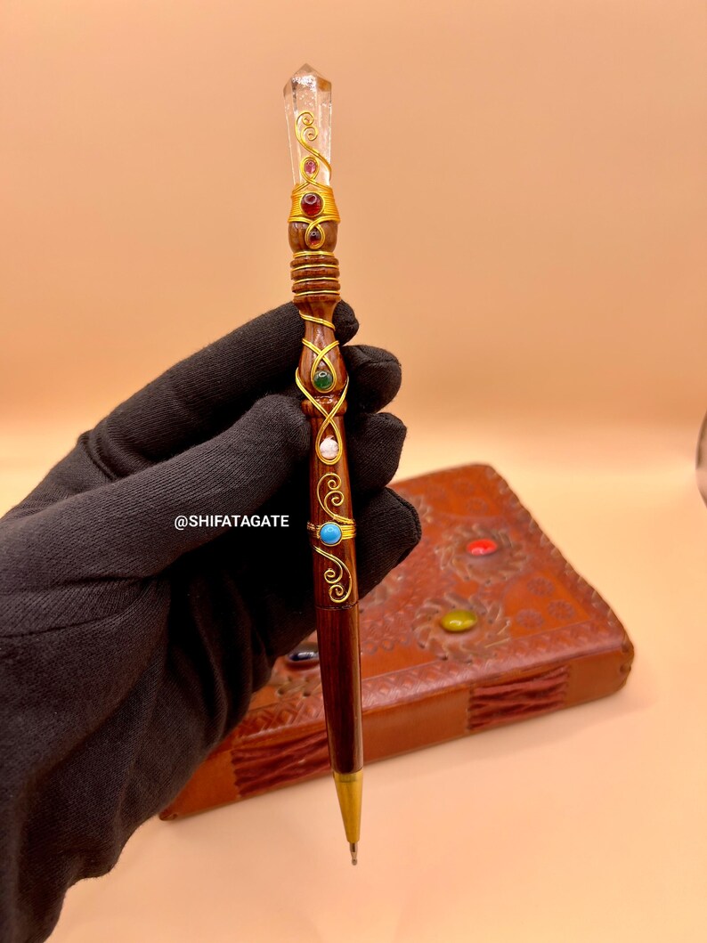 May include: A decorative pen with a clear crystal top and gold accents. The pen features a dark brown wooden body adorned with red, green, and turquoise gemstones. The pen's design includes gold wire wrapping and a gold-colored tip.