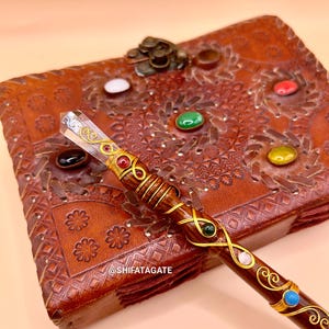May include: A handcrafted wooden wand with a clear crystal tip and decorative gold wire wrapping. The wand is adorned with various colorful gemstones and rests on a brown leather journal with embossed floral patterns. The text "@SHIFATAGATE" is visible.