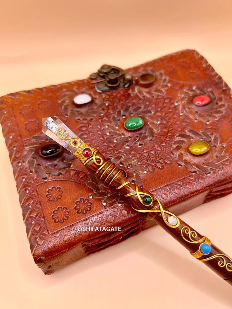 May include: A handcrafted wooden wand with a clear crystal tip and decorative gold wire wrapping. The wand is adorned with various colorful gemstones and rests on a brown leather journal with embossed floral patterns. The text "@SHIFATAGATE" is visible.