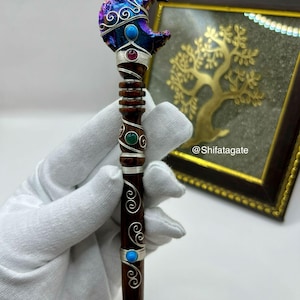 May include: A handcrafted wooden wand with a crescent moon-shaped top. The moon is adorned with iridescent purple and blue accents, silver wire detailing, and turquoise and ruby-colored stones. The wand's handle is dark brown with silver wire accents and turquoise and green stones.