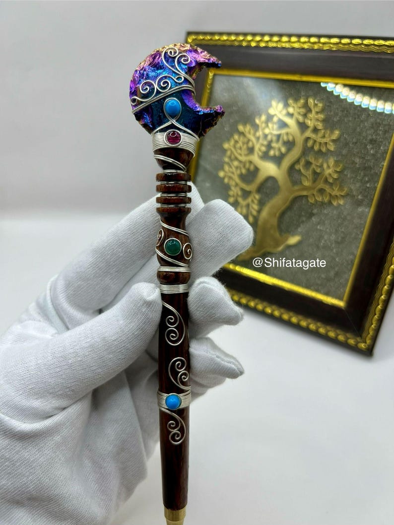 May include: A handcrafted wooden wand with a crescent moon-shaped top. The moon is adorned with iridescent purple and blue accents, silver wire detailing, and turquoise and ruby-colored stones. The wand's handle is dark brown with silver wire accents and turquoise and green stones.