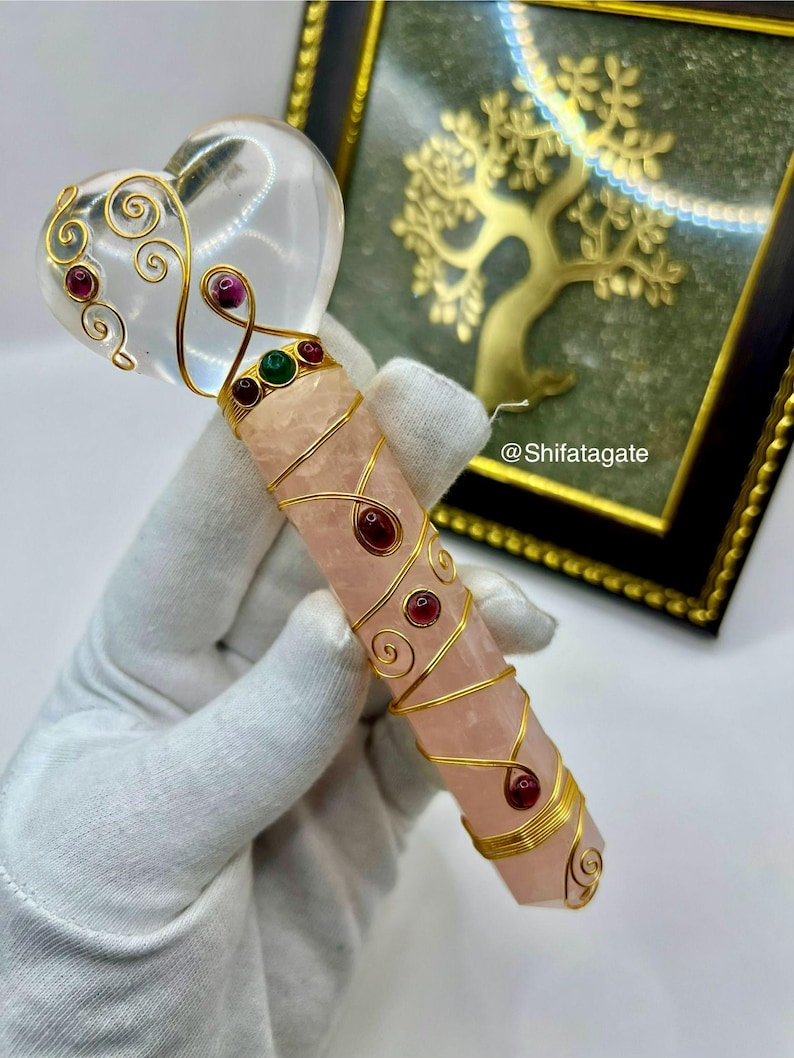 Op de afbeelding: Een kristallen toverstaf met een heldere hartvormige top en een roze rozenkwarts handvat. De toverstaf is versierd met goudkleurige draad en rode en groene edelstenen. De handgreep is omwikkeld met goudkleurige draad met rode edelstenen. De toverstaf wordt vastgehouden door een gehandschoende hand.