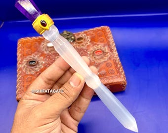May include: A clear, pointed wand with a purple crystal top and gold-coloured accents. The wand is held against a blue background, with a brown, ornate box visible. The wand is approximately 20 cm long.