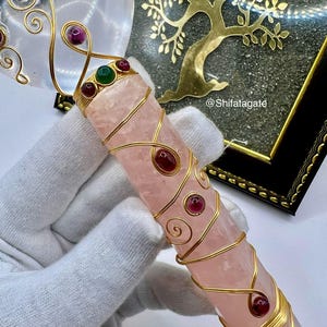 Op de afbeelding: Een roze kristallen toverstaf met gouden draadaccenten en rode en groene edelstenen. De toverstaf wordt in een gehandschoende hand gehouden. Een helder hartvormig kristal en een gouden levensboomkunstwerk staan op de achtergrond.