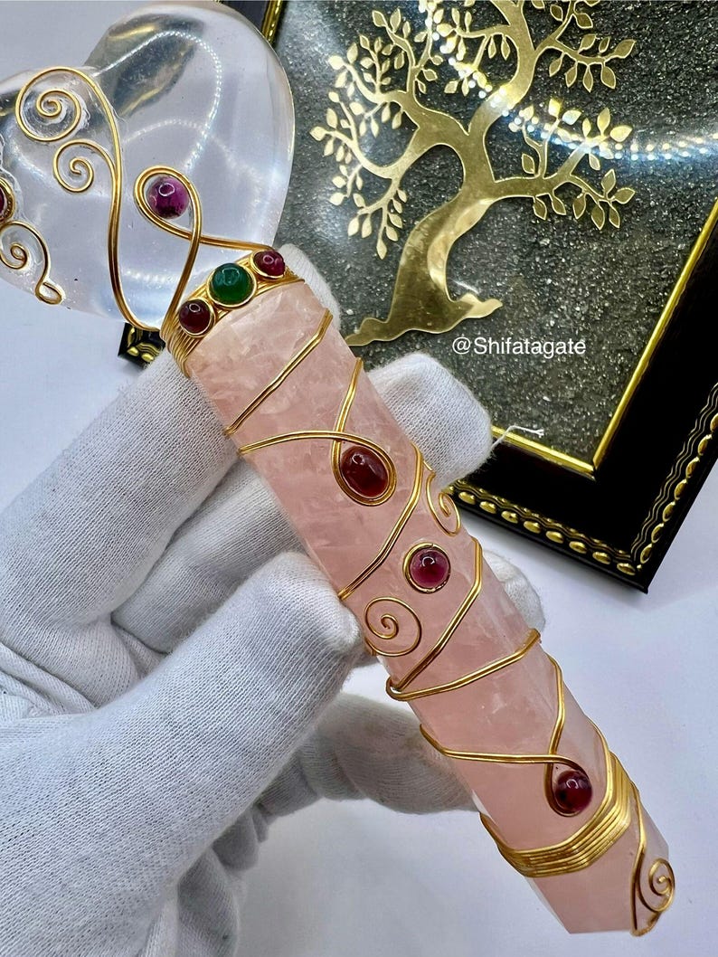 Op de afbeelding: Een roze kristallen toverstaf met gouden draadaccenten en rode en groene edelstenen. De toverstaf wordt in een gehandschoende hand gehouden. Een helder hartvormig kristal en een gouden levensboomkunstwerk staan op de achtergrond.