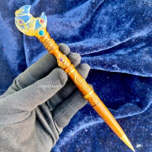 Op de afbeelding: Een handgemaakte houten toverstaf met ingewikkelde gouddraad details. De toverstaf heeft een maanvormige kristallen top met edelstenen en een gouden punt. Een uniek, decoratief item.