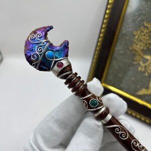 May include: A handcrafted wand with a crescent-shaped, iridescent crystal head. The wand features silver wire detailing, turquoise and ruby-colored accents, and a dark brown wooden handle. Decorative swirls adorn the handle.