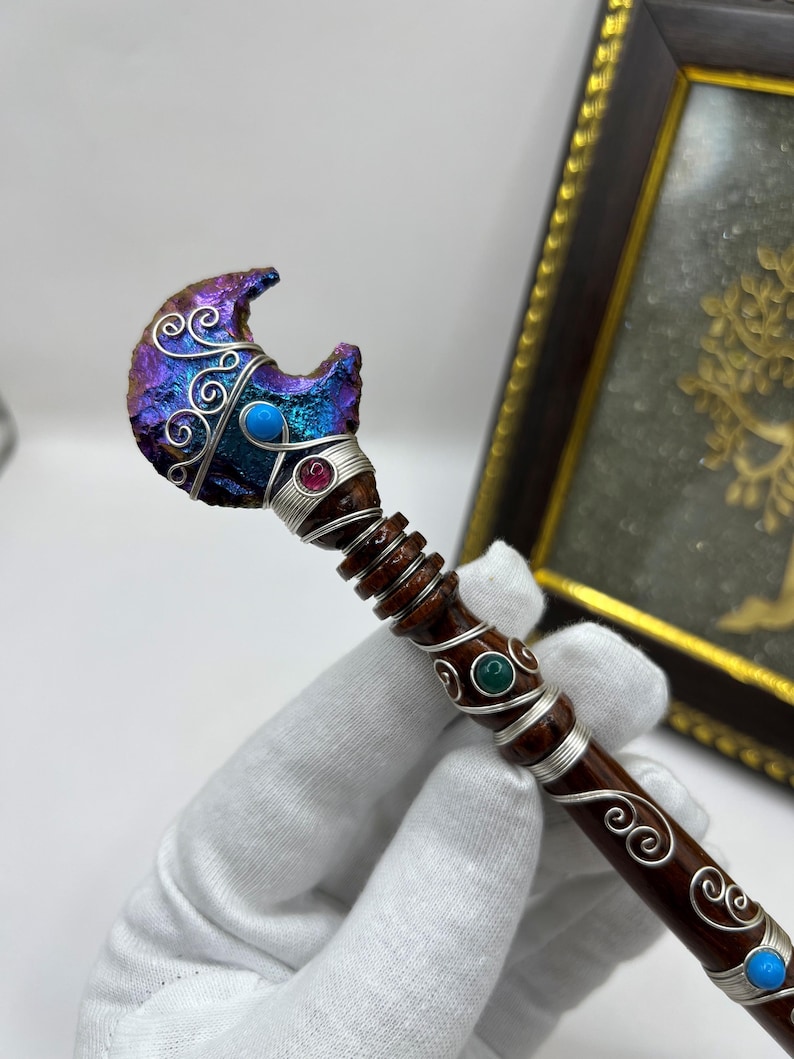 May include: A handcrafted wand with a crescent-shaped, iridescent crystal head. The wand features silver wire detailing, turquoise and ruby-colored accents, and a dark brown wooden handle. Decorative swirls adorn the handle.