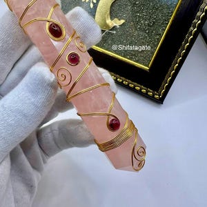Op de afbeelding: Een roze kristallen toverstaf, ongeveer 15 cm lang, omwikkeld met goudkleurig draad met rode en groene edelsteenaccenten. De kristal heeft een facetgeslepen uiteinde. De tekst "@Shifatagate" is zichtbaar.