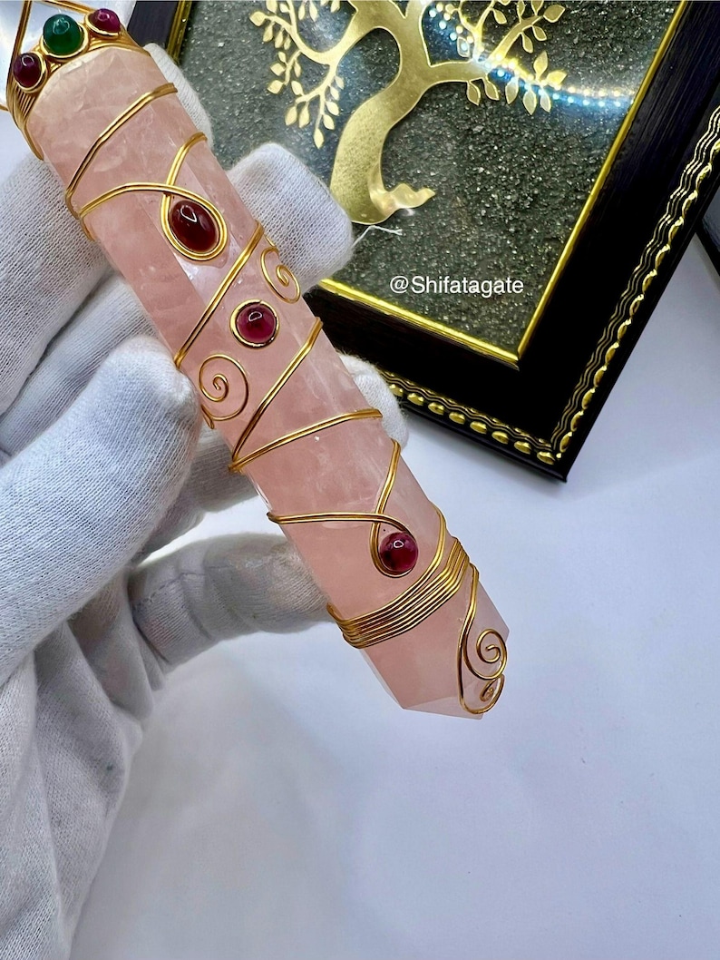 Op de afbeelding: Een roze kristallen toverstaf, ongeveer 15 cm lang, omwikkeld met goudkleurig draad met rode en groene edelsteenaccenten. De kristal heeft een facetgeslepen uiteinde. De tekst "@Shifatagate" is zichtbaar.