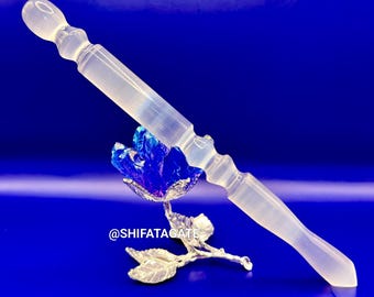 May include: A clear, elongated crystal wand with rounded ends, resting on a silver-colored rose with a blue crystal center. The background is a solid, deep blue. The wand is approximately 6 inches long.