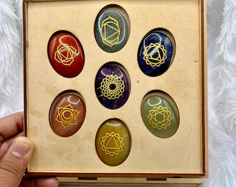 May include: A set of seven oval-shaped polished stones in a light-colored wooden display box. Each stone features a unique gold-colored engraved symbol. The stones are arranged in a circular pattern with colors including red, dark green, blue, purple, light green, yellow, and brown.