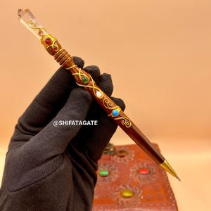 May include: A decorative pen with a clear crystal top and a dark brown wooden body. The pen is adorned with gold-colored filigree and colorful gemstones. The pen has a gold-colored tip. The text "@SHIFATAGATE" is visible.