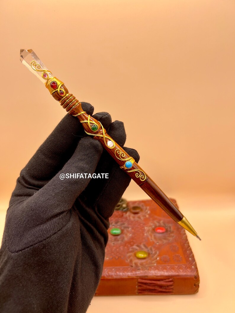 May include: A decorative pen with a clear crystal top and a dark brown wooden body. The pen is adorned with gold-colored filigree and colorful gemstones. The pen has a gold-colored tip. The text "@SHIFATAGATE" is visible.