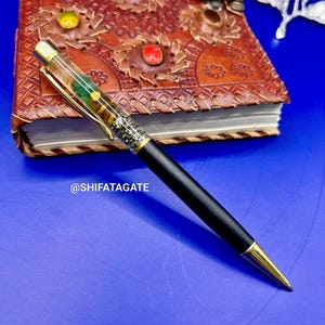 K&ouml;nnte beinhalten: Ein schwarzer Stift mit goldenen Akzenten und einem dekorativen, mit Edelsteinen gef&uuml;llten Schaft liegt auf einem braunen Lederjournal mit farbenfrohen Edelsteinverzierungen. Der Stift hat ein schlankes Design und eine goldfarbene Spitze. Der Text "@SHIFATAGATE" ist sichtbar.
