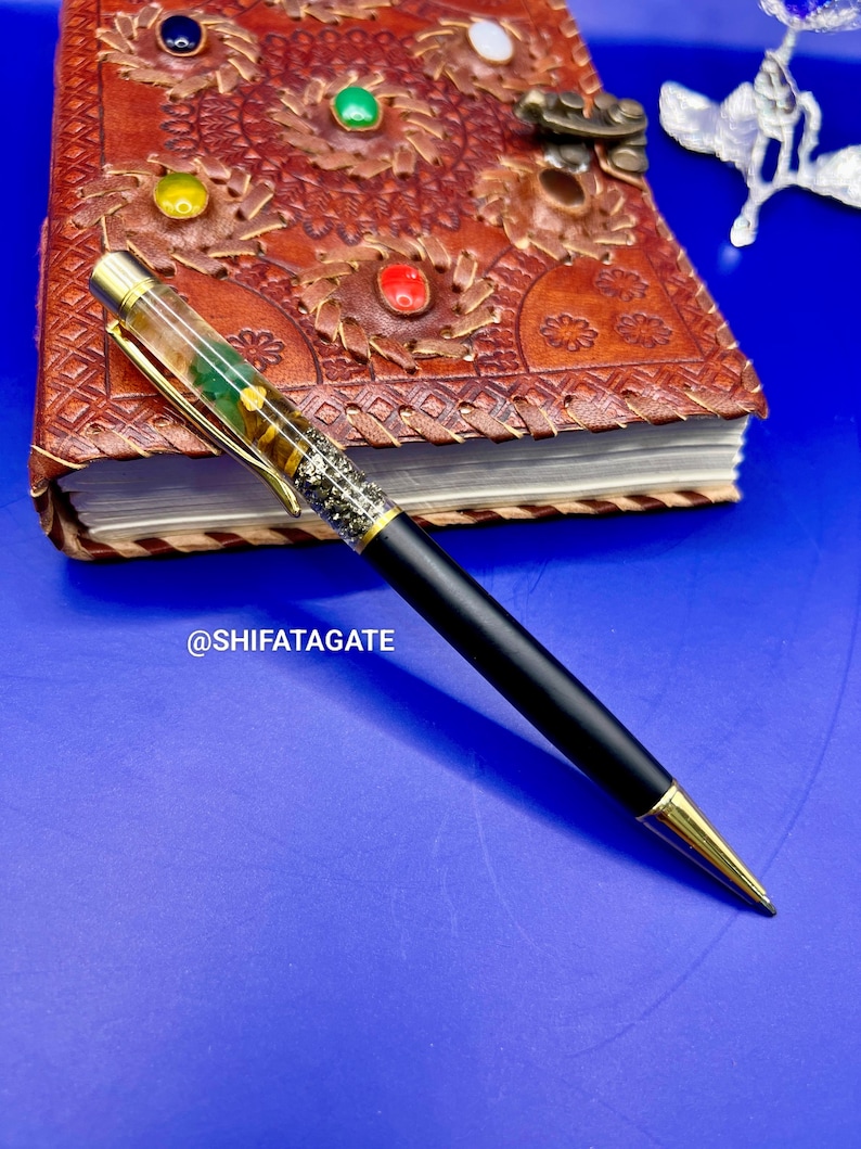 K&ouml;nnte beinhalten: Ein schwarzer Stift mit goldenen Akzenten und einem dekorativen, mit Edelsteinen gef&uuml;llten Schaft liegt auf einem braunen Lederjournal mit farbenfrohen Edelsteinverzierungen. Der Stift hat ein schlankes Design und eine goldfarbene Spitze. Der Text "@SHIFATAGATE" ist sichtbar.