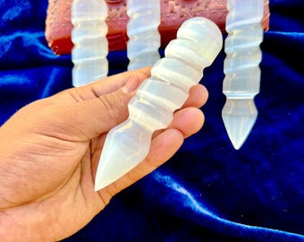 May include: A hand holds a white, spiraled crystal wand with a pointed tip. Several similar wands are arranged in the background, along with a decorative, pink, jeweled box. The background is a dark blue velvet.