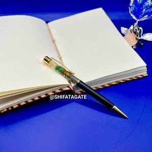K&ouml;nnte beinhalten: Ein offenes Notizbuch mit leeren Seiten und einem schwarzen Stift mit goldenen Akzenten. Der Stift hat einen transparenten Schaft, der mit gr&uuml;nen und silbernen Glitzer gef&uuml;llt ist. Ein blauer Hintergrund und eine blaue Metallblume sind ebenfalls zu sehen.