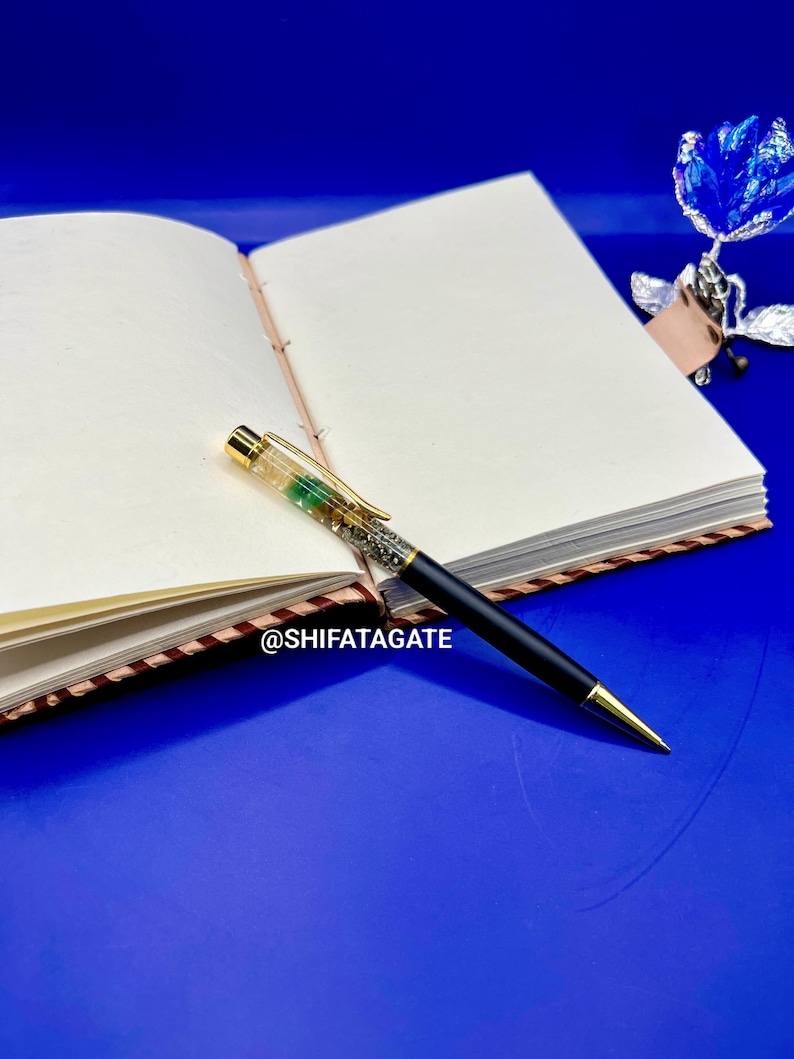 K&ouml;nnte beinhalten: Ein offenes Notizbuch mit leeren Seiten und einem schwarzen Stift mit goldenen Akzenten. Der Stift hat einen transparenten Schaft, der mit gr&uuml;nen und silbernen Glitzer gef&uuml;llt ist. Ein blauer Hintergrund und eine blaue Metallblume sind ebenfalls zu sehen.