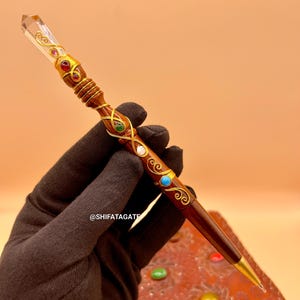 May include: A handcrafted wooden pen with a clear crystal top and gold accents. The pen is adorned with colorful gemstones and intricate gold wire detailing. The pen has a gold nib and is resting on a leather-bound journal. The text "@SHIFATAGATE" is visible.