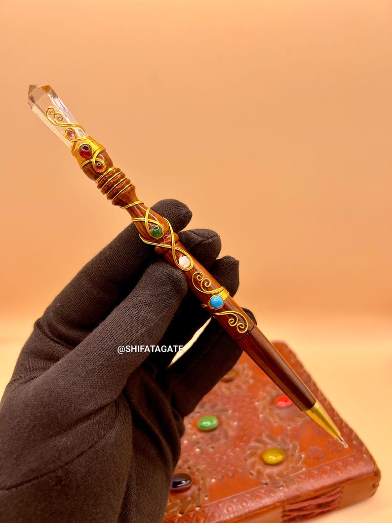 May include: A handcrafted wooden pen with a clear crystal top and gold accents. The pen is adorned with colorful gemstones and intricate gold wire detailing. The pen has a gold nib and is resting on a leather-bound journal. The text "@SHIFATAGATE" is visible.