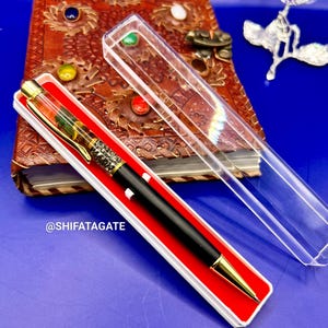 K&ouml;nnte beinhalten: Ein schwarzer und goldener Stift in einer rot-wei&szlig;en Schachtel, mit einem detaillierten Design am Stiftk&ouml;rper. Der Stift liegt vor einem ledergebundenen Notizbuch mit farbigen Edelsteinen, vor blauem Hintergrund. Der Text "@SHIFATAGATE" ist sichtbar.