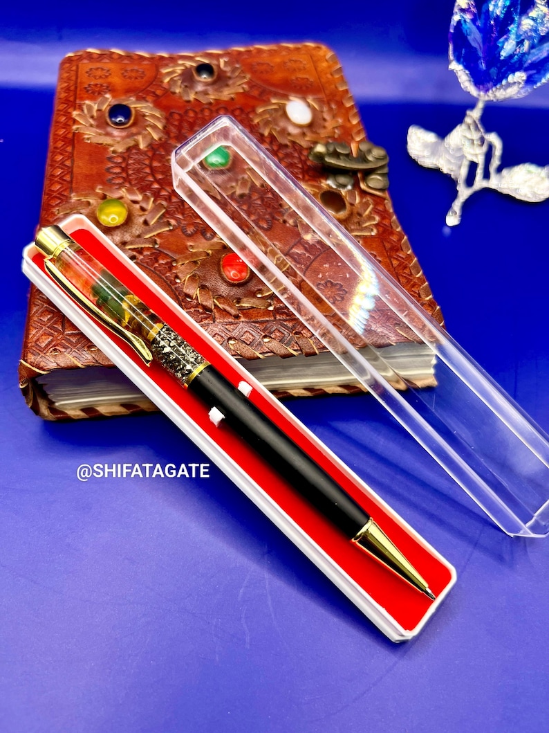 K&ouml;nnte beinhalten: Ein schwarzer und goldener Stift in einer rot-wei&szlig;en Schachtel, mit einem detaillierten Design am Stiftk&ouml;rper. Der Stift liegt vor einem ledergebundenen Notizbuch mit farbigen Edelsteinen, vor blauem Hintergrund. Der Text "@SHIFATAGATE" ist sichtbar.