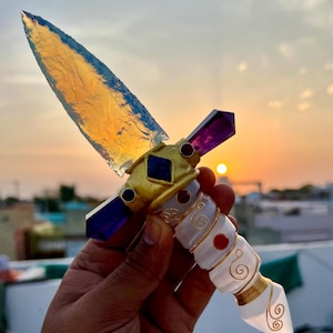 May include: A decorative dagger with a translucent blade and a handle adorned with purple and red gemstones, gold accents, and white detailing. The blade is a light amber color. The dagger is held up against a sunset backdrop.