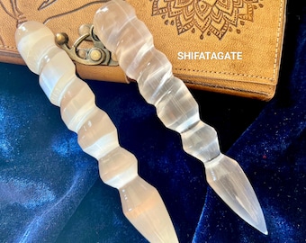 May include: Two white crystal wands with a textured, spiraled design. The wands are lying on a blue velvet surface. The text "SHIFATAGATE" is visible on a brown leather surface in the background.