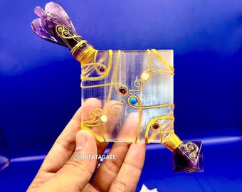 May include: A clear, rectangular crystal decorated with gold wire and small gemstones. The ends have purple amethyst accents. The text "@SHIFATAGATE" is visible. The object is held against a blue background.