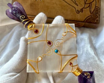 May include: A clear crystal wand with gold wire wrapped around it and various coloured gemstones. The wand is held in a white gloved hand. The wand is topped with a purple amethyst crystal.