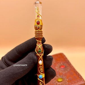 May include: A handcrafted wooden wand with a clear crystal tip and gold wire accents. The wand is adorned with red, green, white, and blue gemstones. The wand has a dark brown finish and is held by a gloved hand.