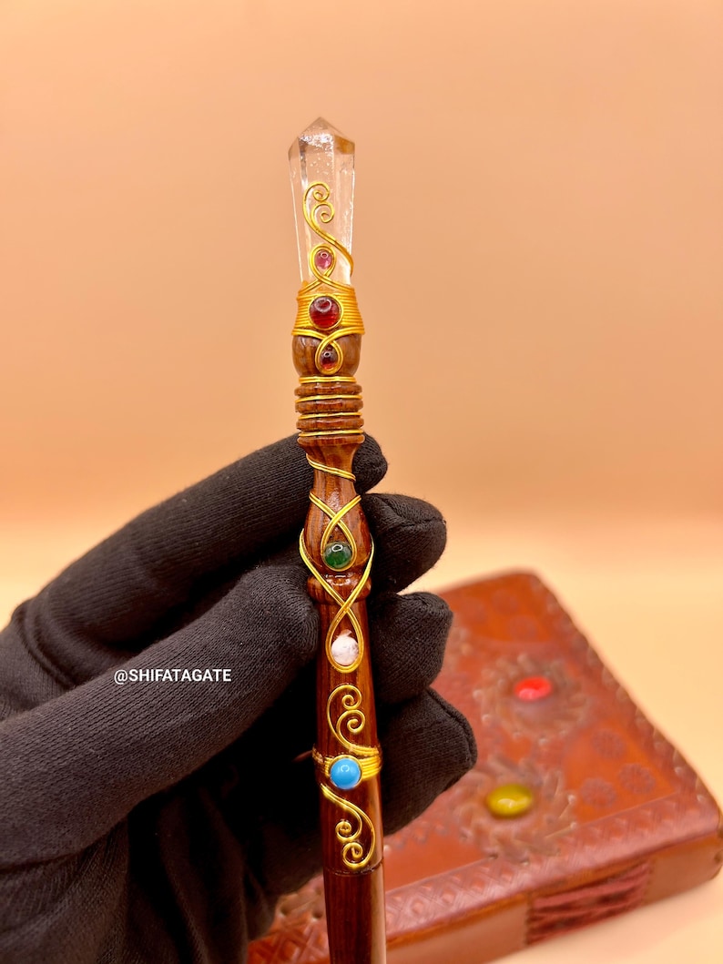 May include: A handcrafted wooden wand with a clear crystal tip and gold wire accents. The wand is adorned with red, green, white, and blue gemstones. The wand has a dark brown finish and is held by a gloved hand.